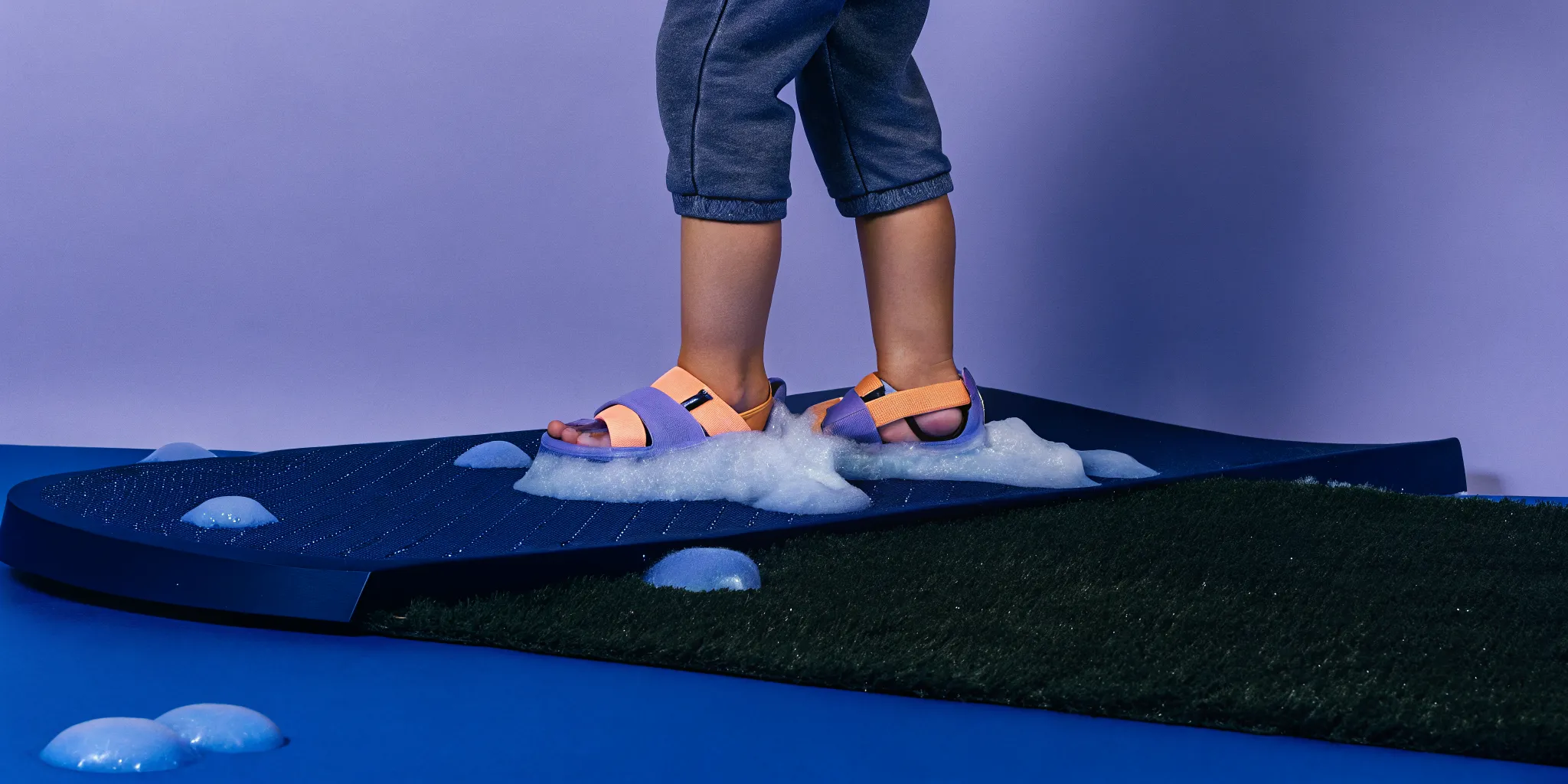 Child wearing water shoes while stepping carefully into a foam play area with non-slip matting on grass for safety