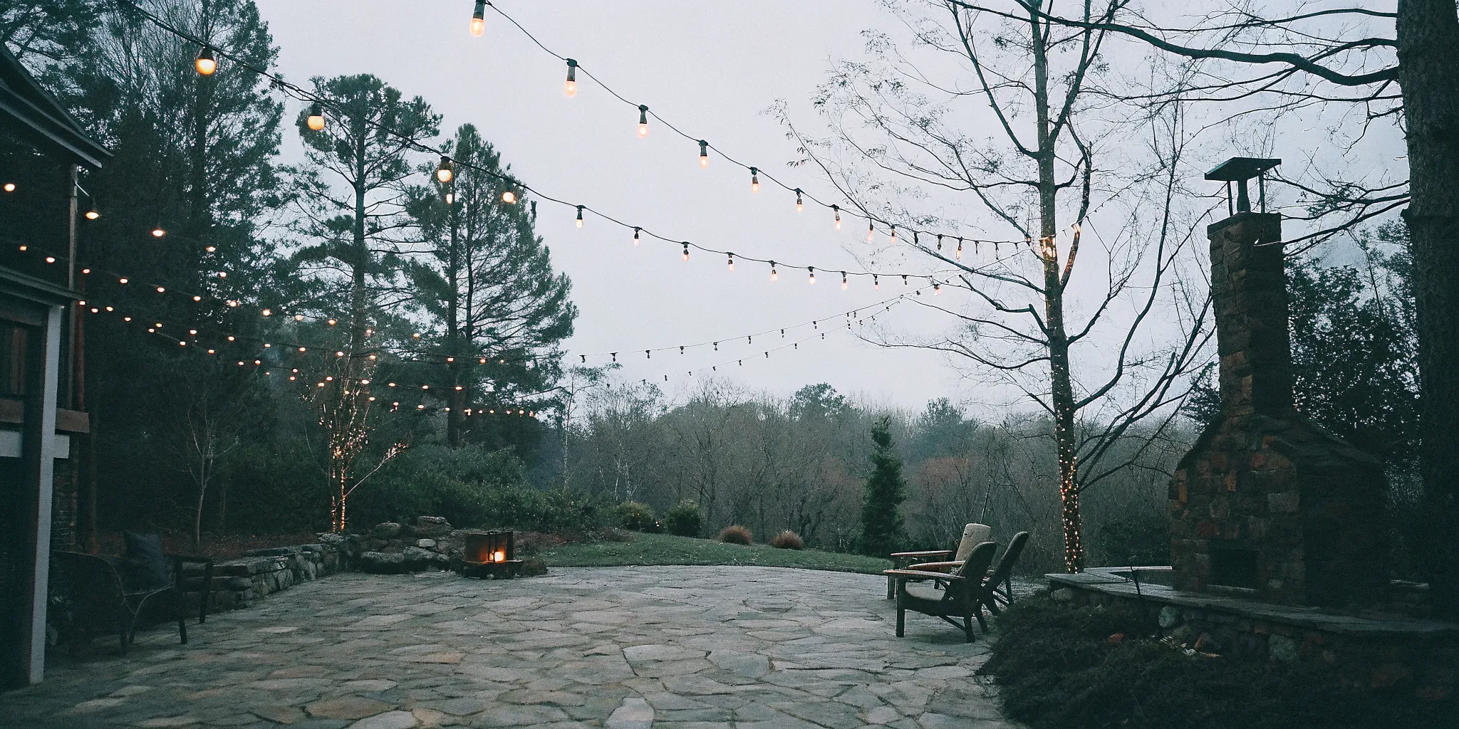 A warm Carolina evening backyard with ambient string lights, low-voltage pathway lighting on a stone patio, and soft accent uplighting on trees and a fire feature