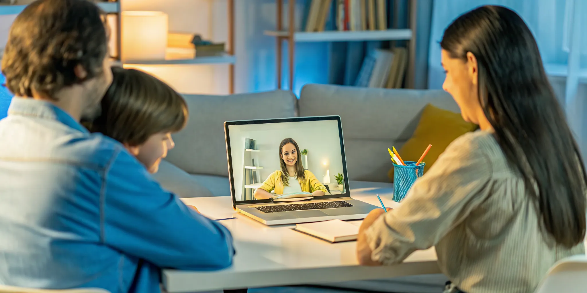 A student, teacher, and parent working together around a home desk during a virtual lesson