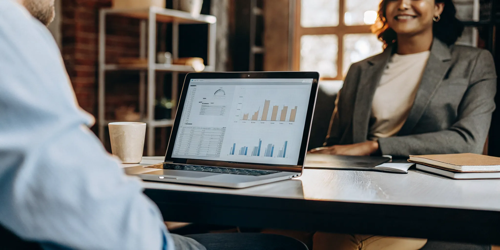 A professional modern office workspace with a laptop showing financial dashboards and a CPA meeting with a small business owner in the background.