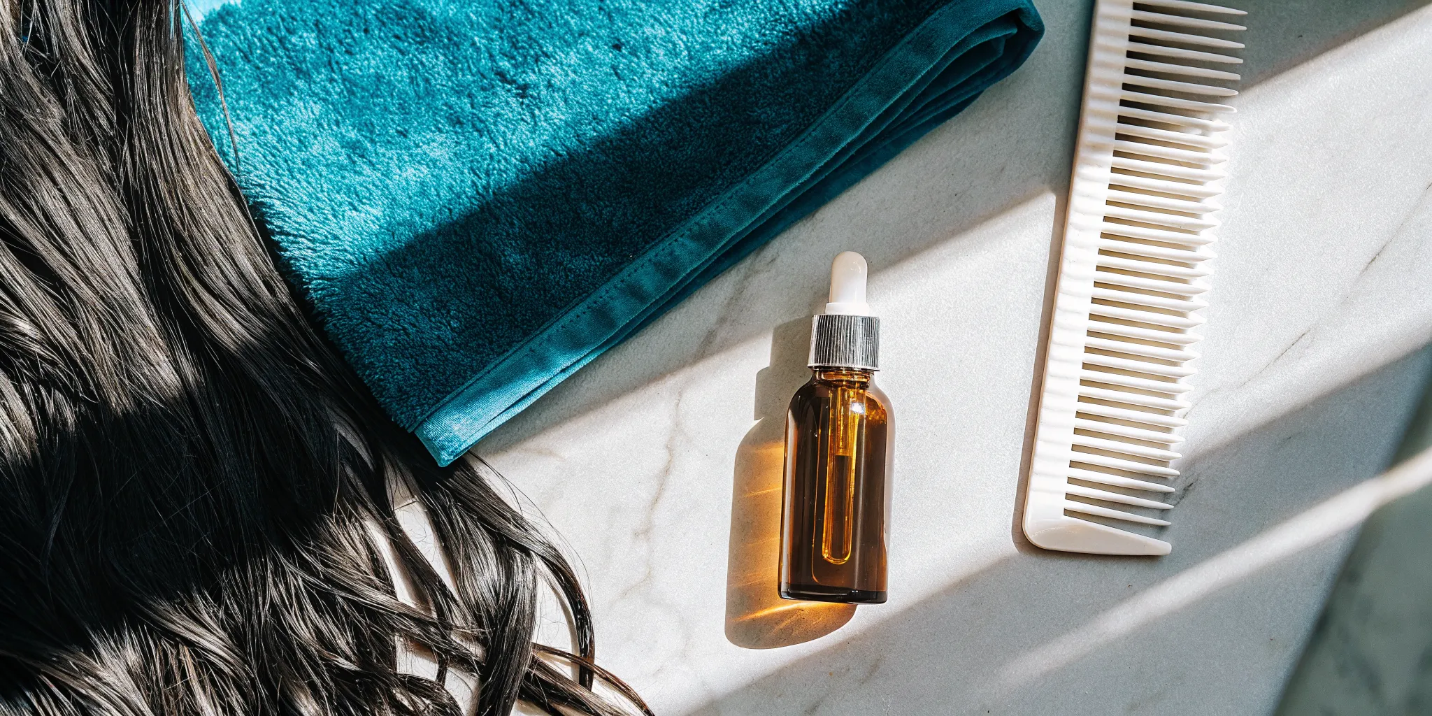 Flat lay of a golden hair oil dropper, comb, and glossy color-treated strands on white marble