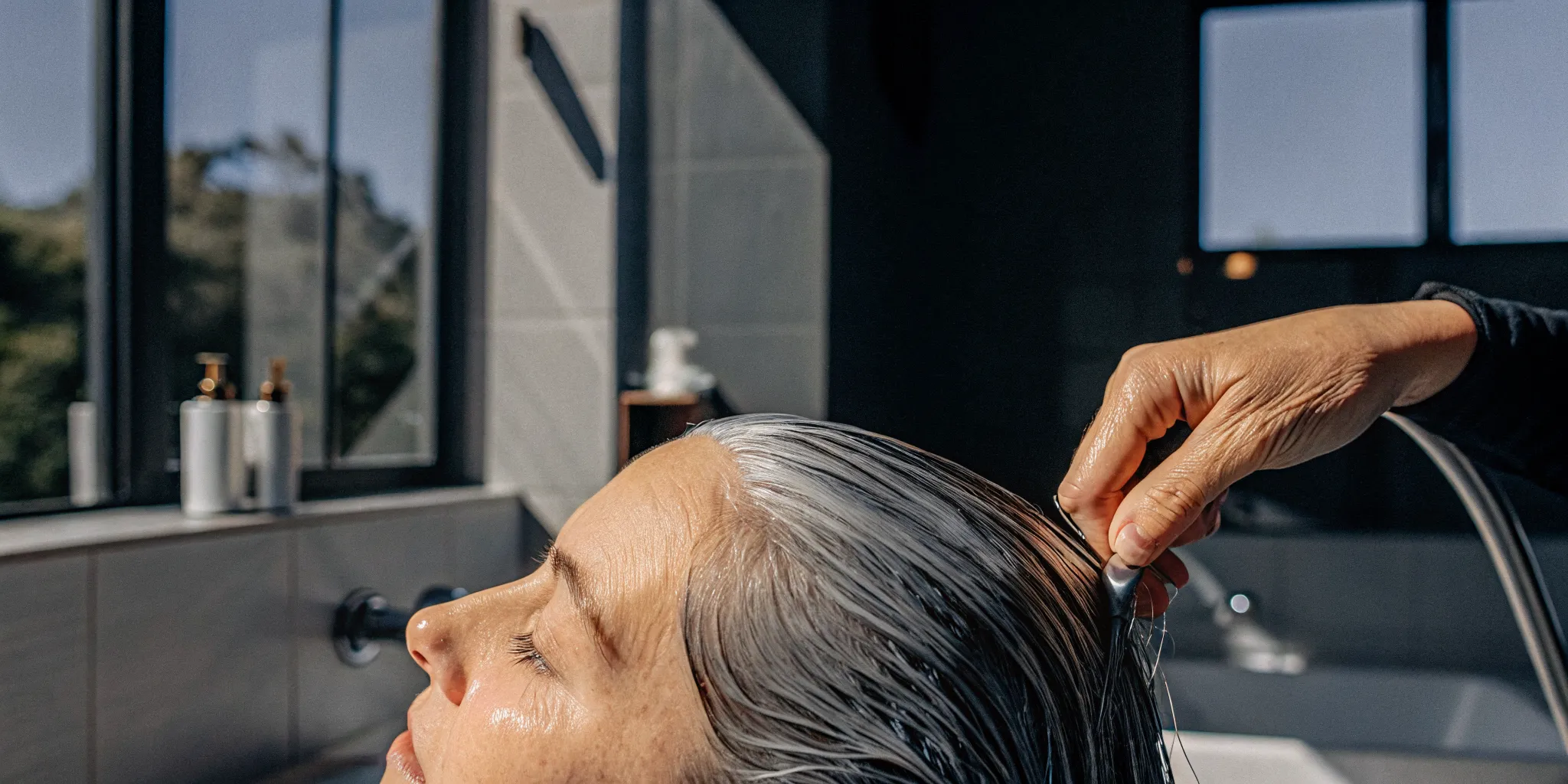 Woman with silver-gray roots receiving a deep conditioning hair treatment in a bright modern bathroom