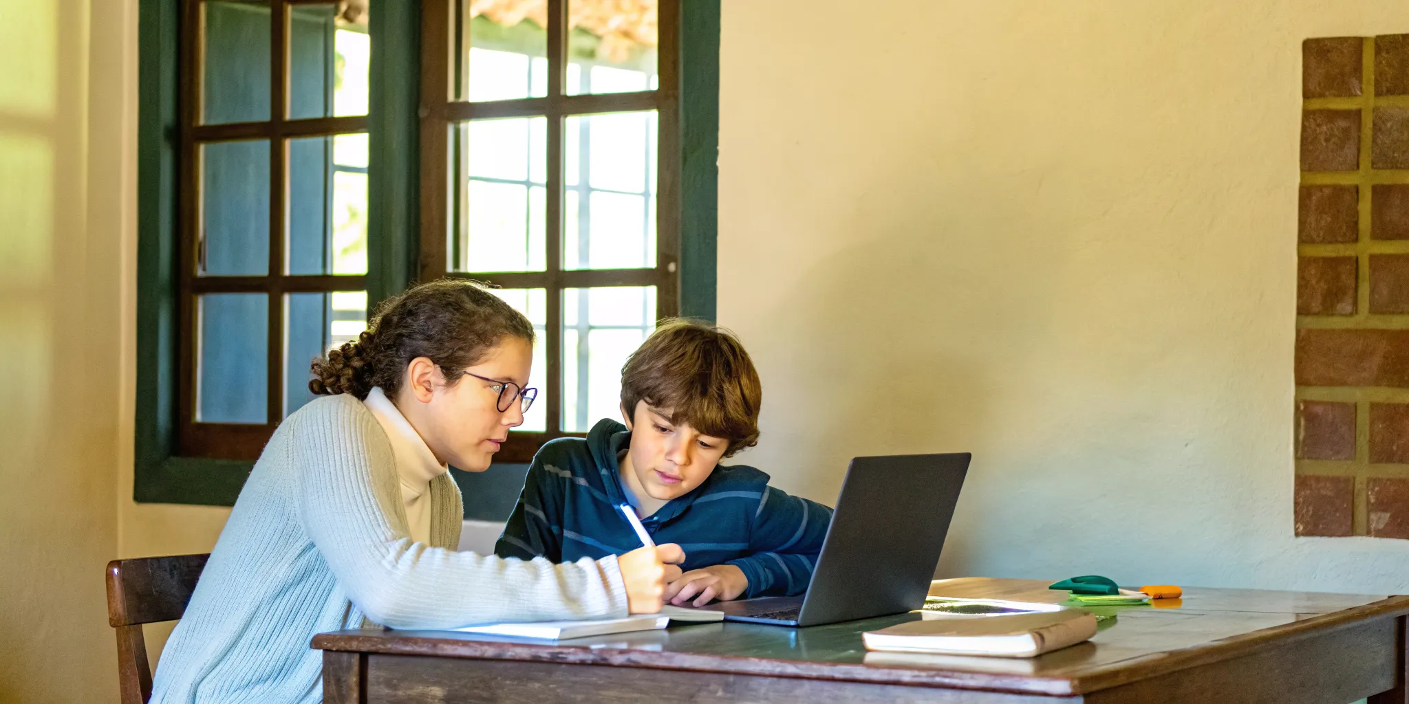 A focused student working on algebra problems with a tutor by their side, notebooks and laptop visible on a warm summer study desk