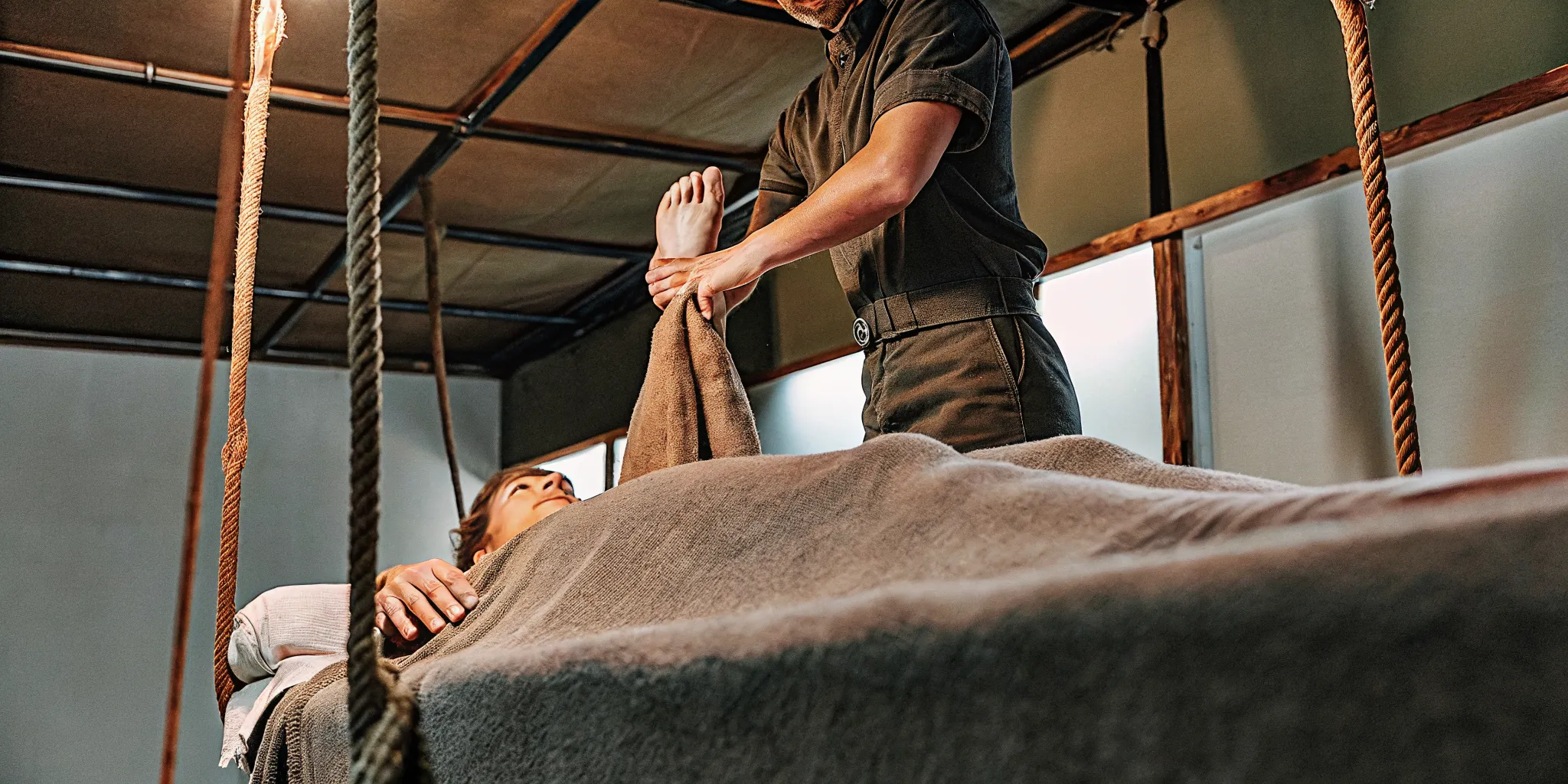 A massage therapist performing barefoot ashiatsu massage while gripping overhead bars