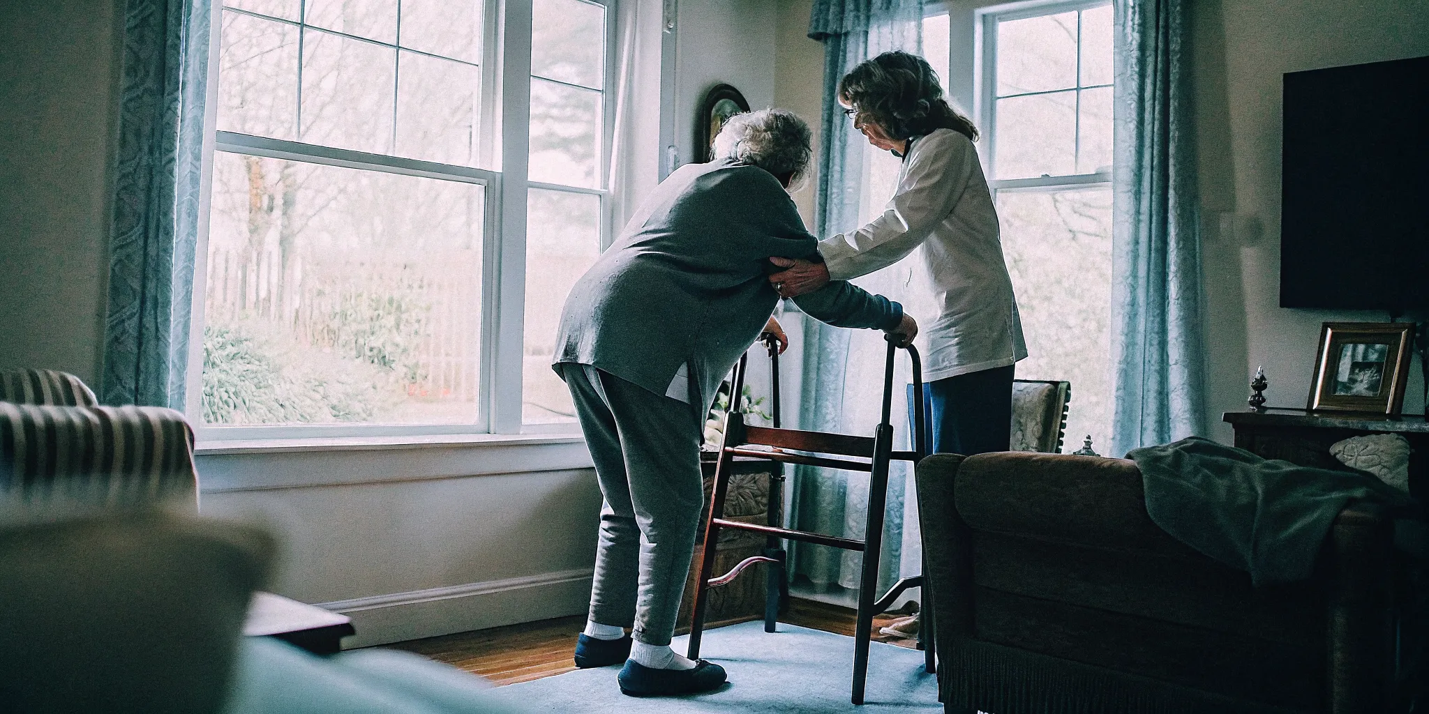 Older adult doing supported balance exercises beside a sturdy chair with a caregiver nearby