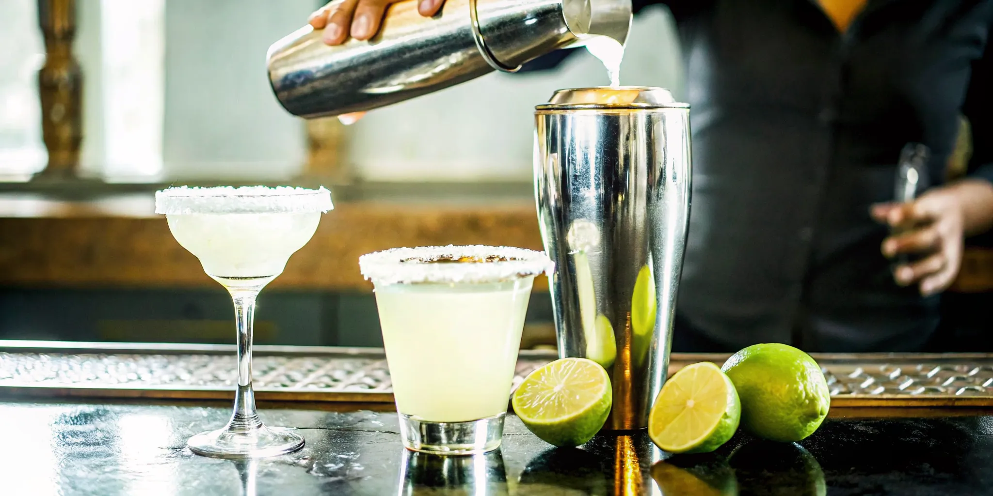 Bartender pouring agave spirit into a cocktail shaker with lime and agave on the bar